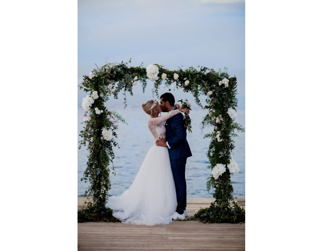 Couple s'embrassant sous une arche de fleur devant la mer.